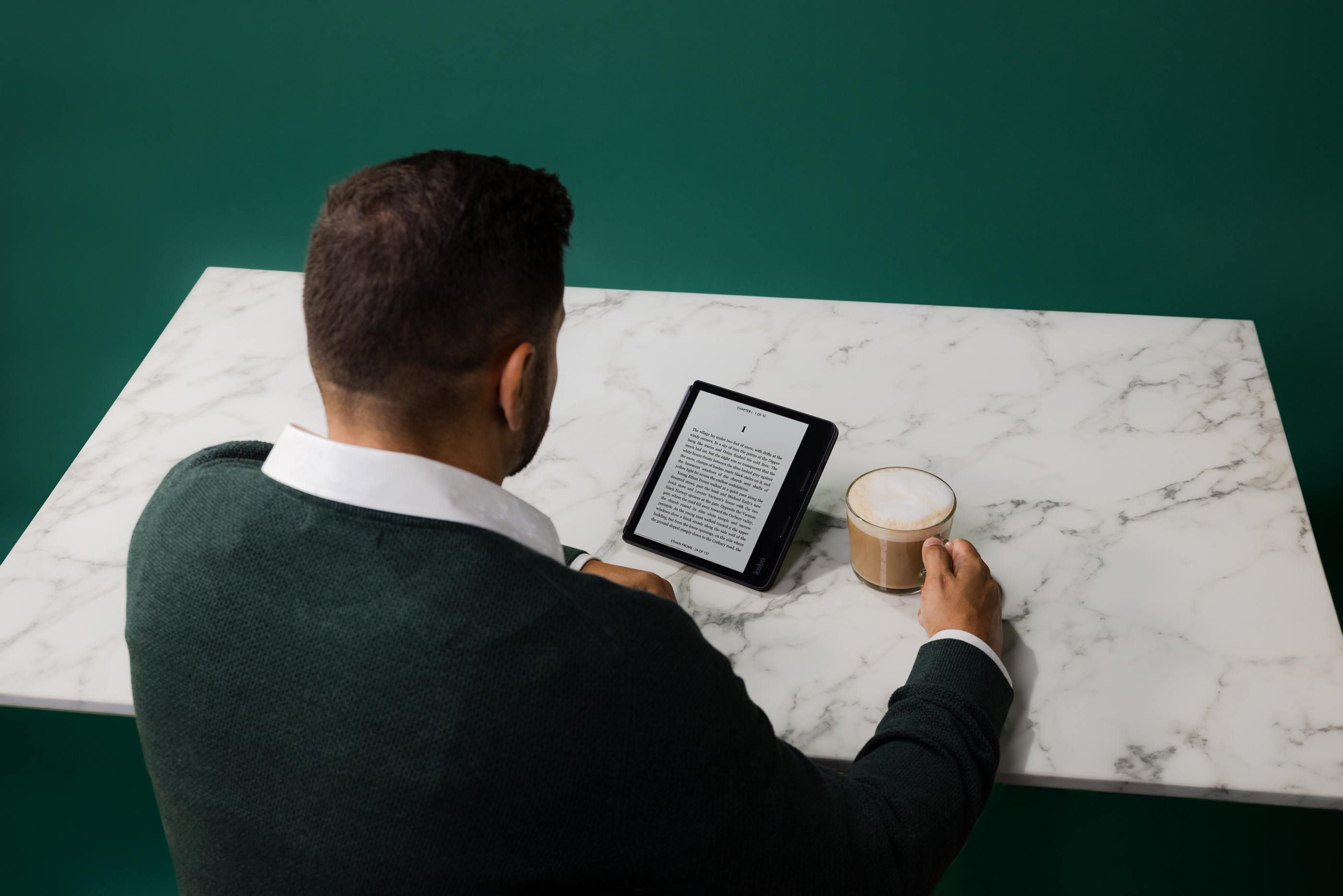 Eine Person liest ein Buch auf einem Tablet und hält eine Kaffeetasse. Der Hintergrund ist ein weißer Marmortisch und eine grüne Wand.