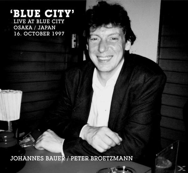 Schwarz-Weiß-Foto eines lächelnden Mannes in einem dunklen Raum. Text: 'BLUE CITY' live at Blue City.