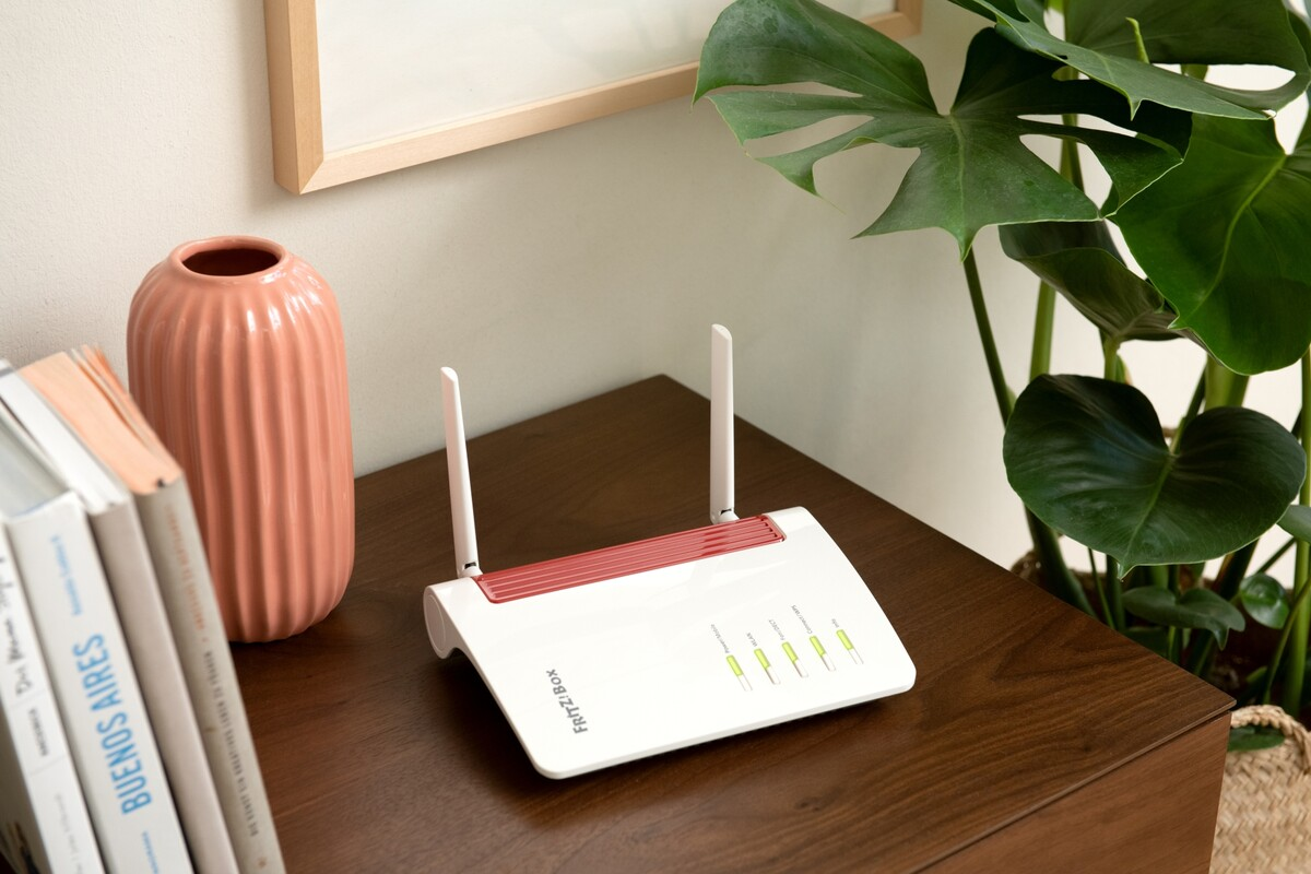 Un router bianco con due antenne e una striscia rossa. Posizionato su un tavolo marrone accanto a un vaso e una pianta.