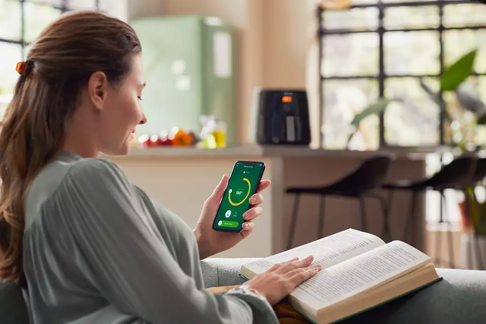 Une femme utilise un téléphone pour contrôler un appareil tout en lisant un livre sur un canapé.