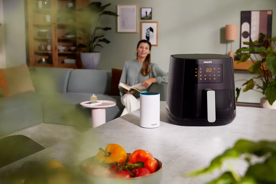 Une friteuse à air sur une table avec une femme lisant et un appareil Amazon Alexa.