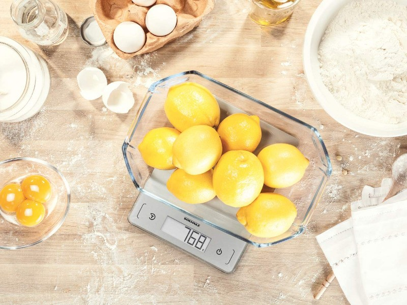 A glass dish with lemons on a scale. Eggs and flour are in the background.