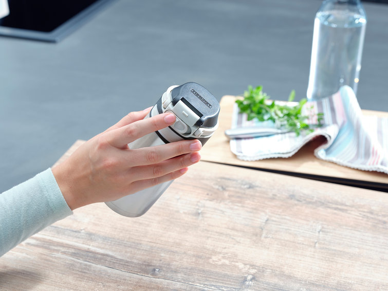 A hand holding a metal water bottle. The lid is black with a white button, the bottle is silver. A table is in the scene.
