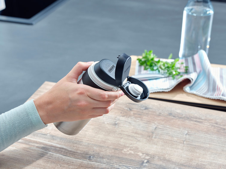 Hand holding a stainless steel and black water bottle. Background: a wooden table and kitchen.