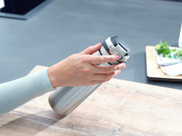 A person's hand holding a silver and black insulated bottle. The background shows a kitchen setting.
