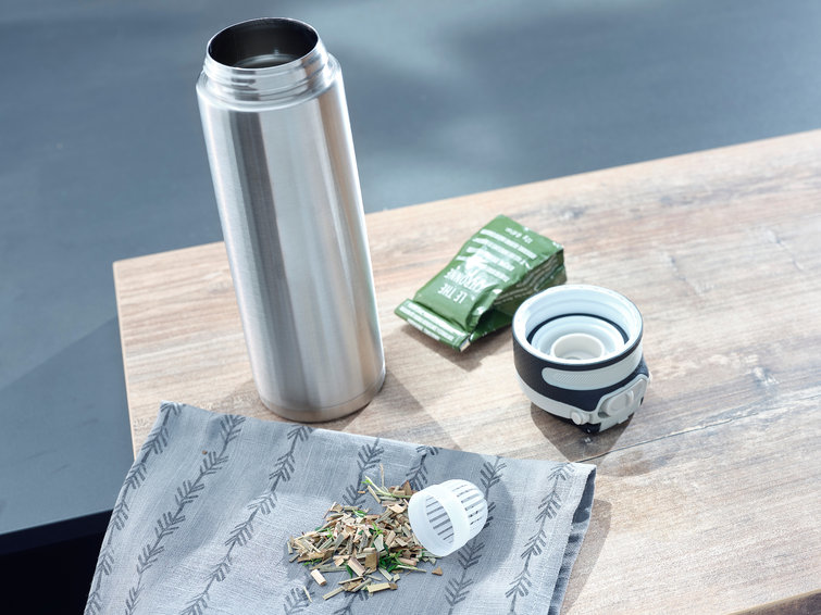 A stainless steel water bottle, a tea bag, and some tea leaves on a wooden table.