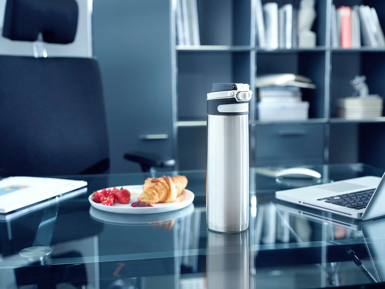Silver water bottle on a glass desk next to a plate of food and a laptop. Office setting.