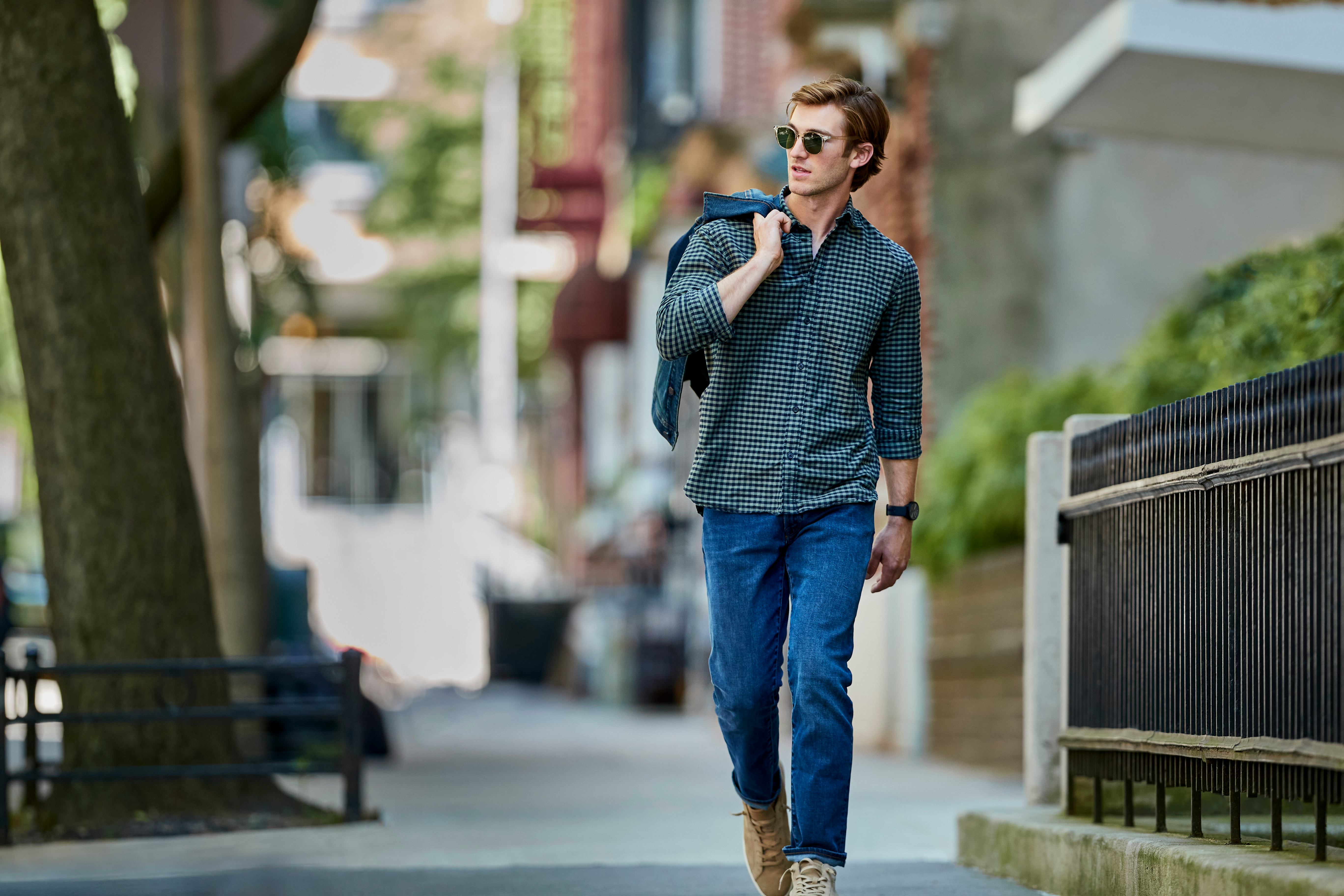 Un homme en jean et chemise marche dans une rue de la ville, avec une veste sur l'épaule.