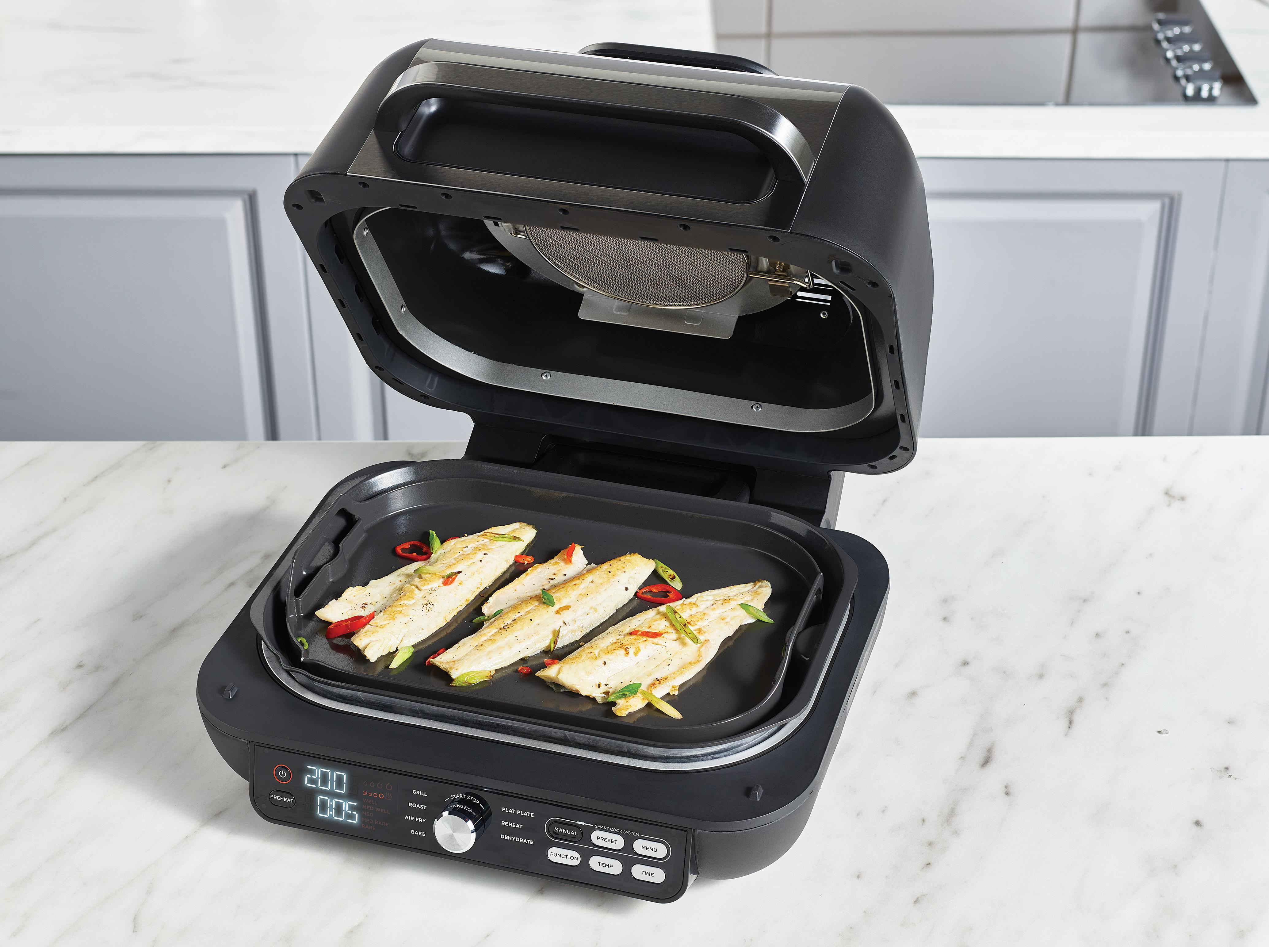Ein schwarzer Grill mit vier Fischfilets, garniert mit Chili und Frühlingszwiebeln.