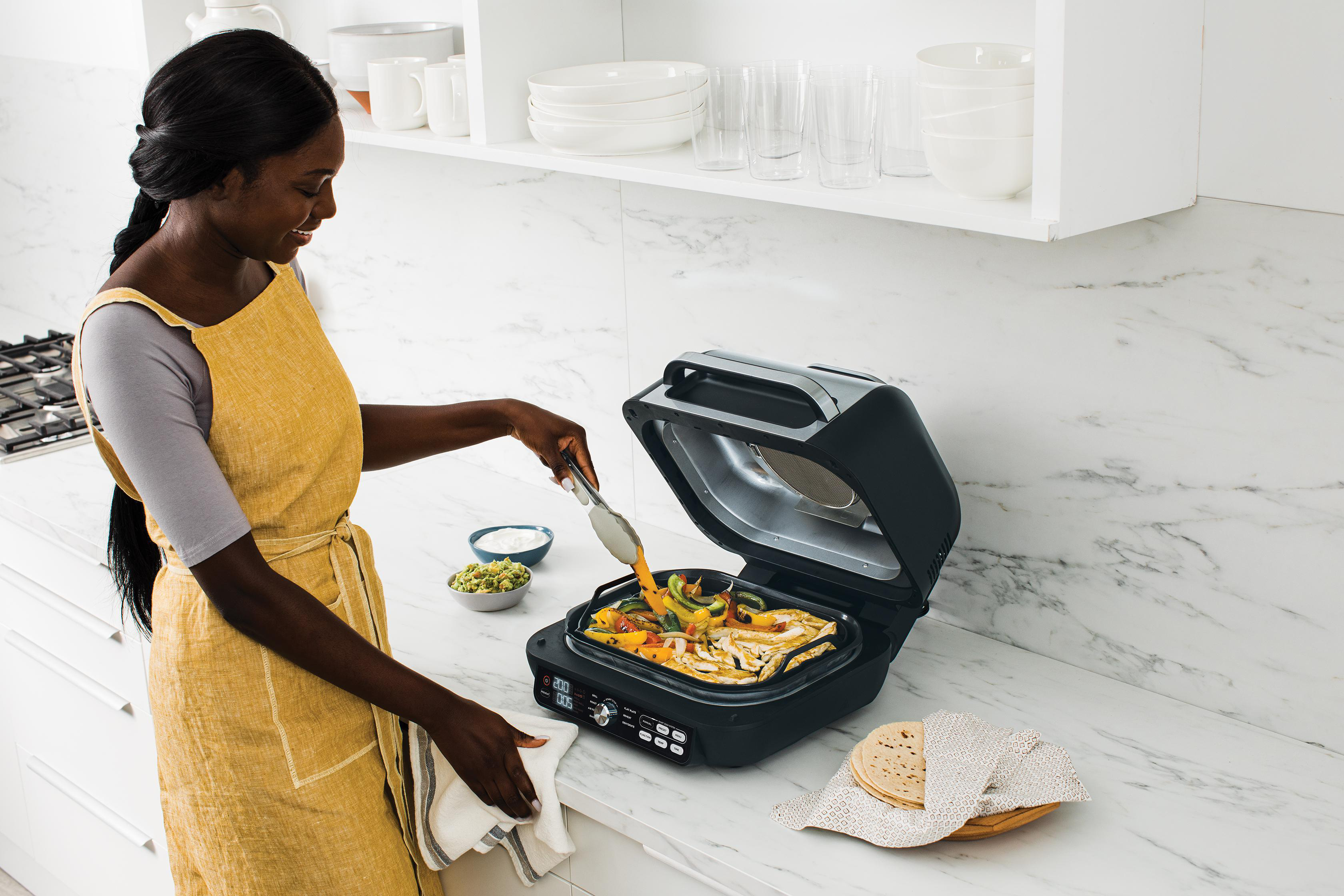 Eine Frau mit gelber Schürze kocht Gemüse auf einem Elektrogrill.