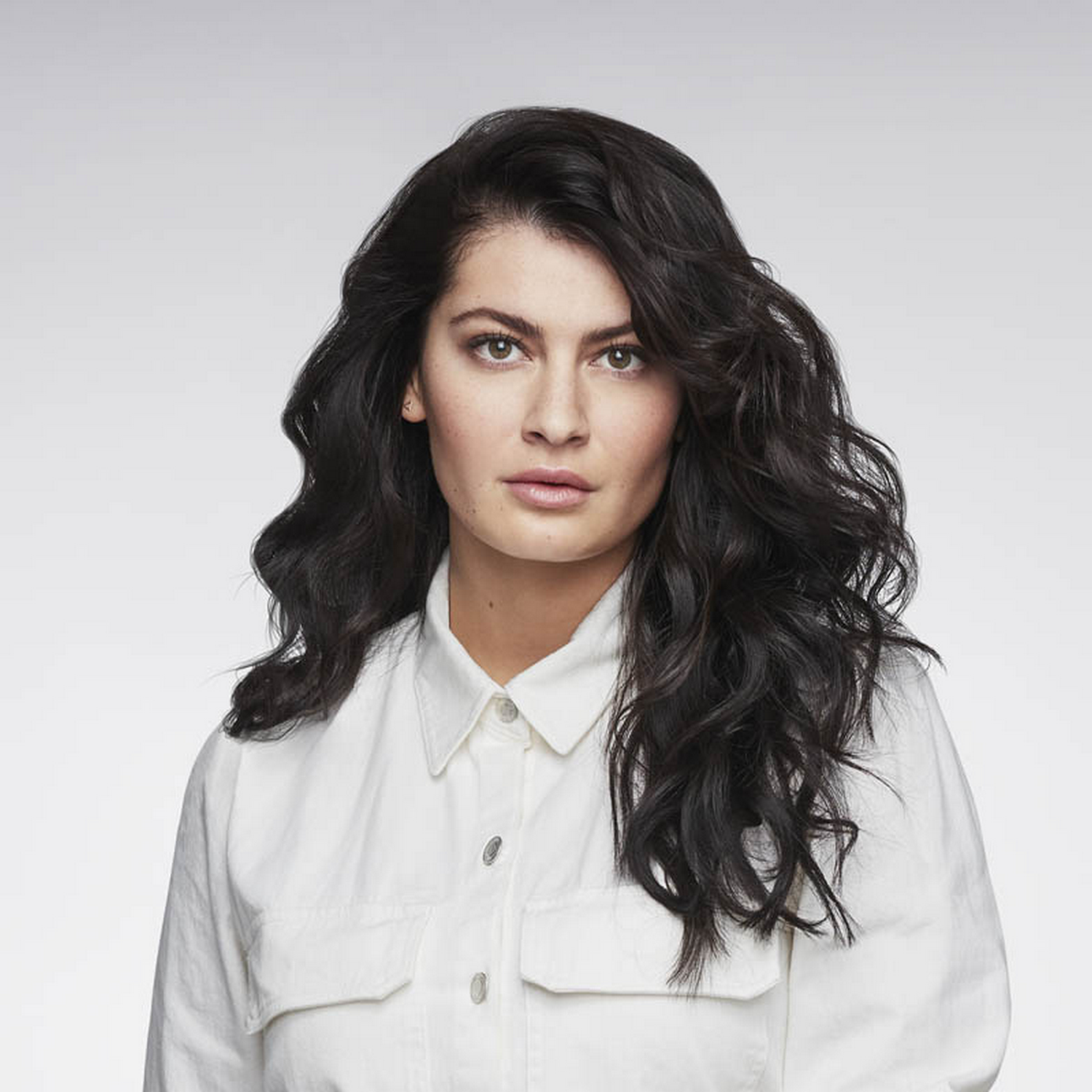 Una mujer con cabello largo, oscuro y ondulado. Lleva una camisa blanca.