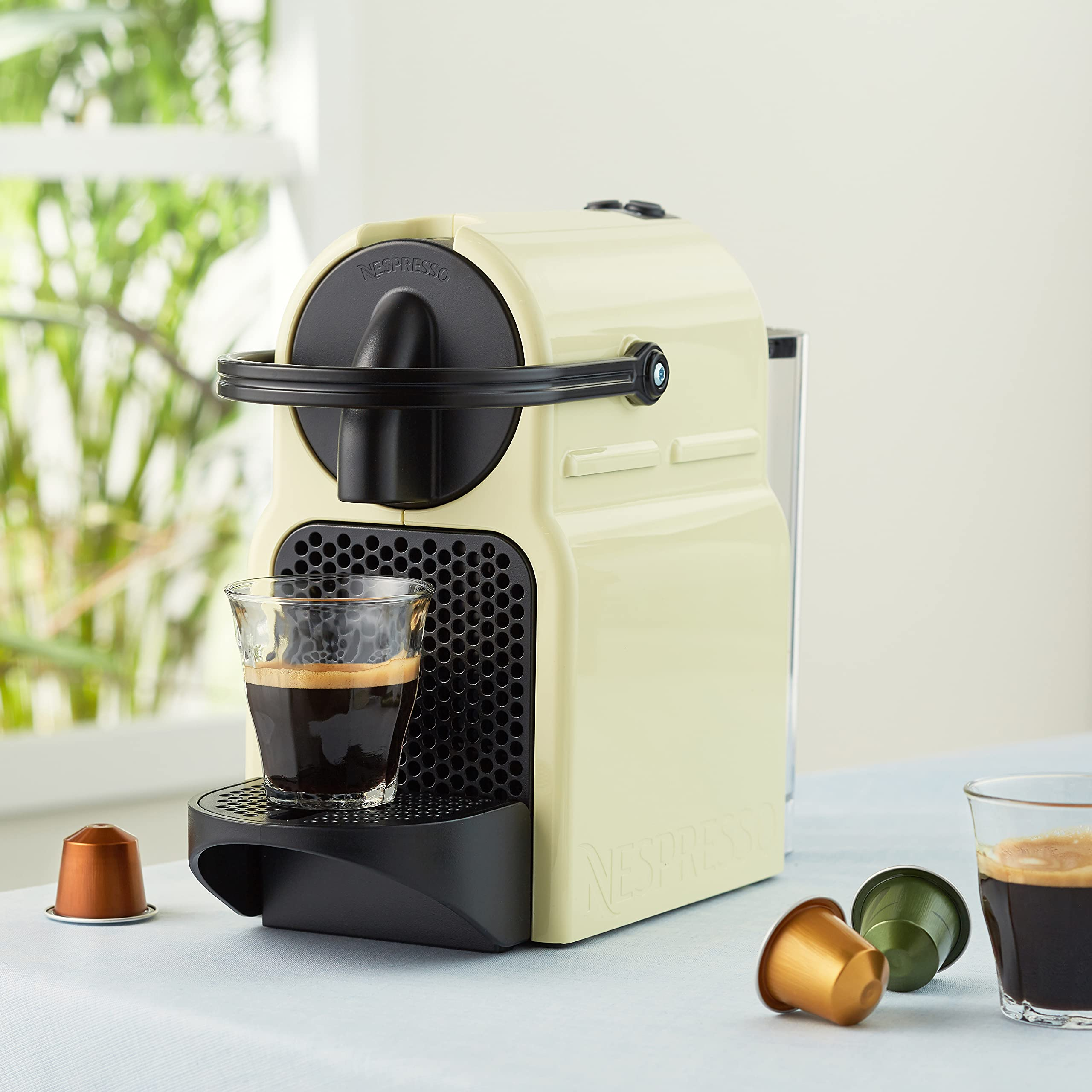 Krémszínű Nespresso gép egy kávézó pohárral és kapszulákkal. Egy ablak látható a háttérben.