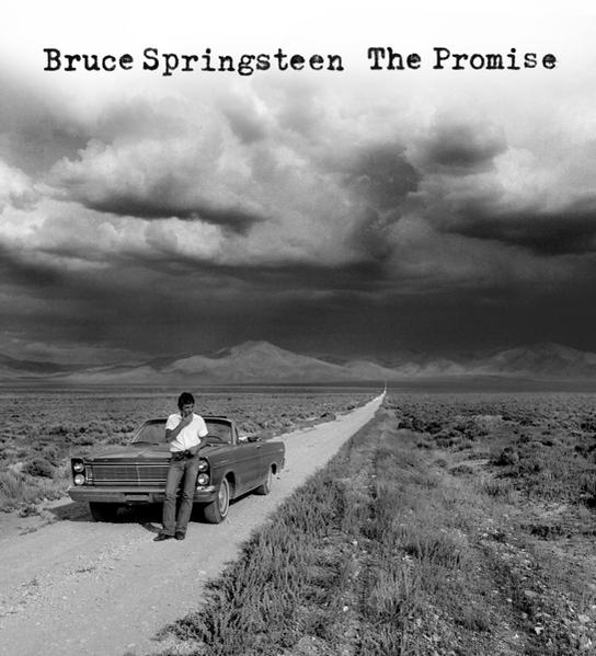 Fekete-fehér fotó Bruce Springsteennel, aki egy autóhoz támaszkodik egy hosszú úton.