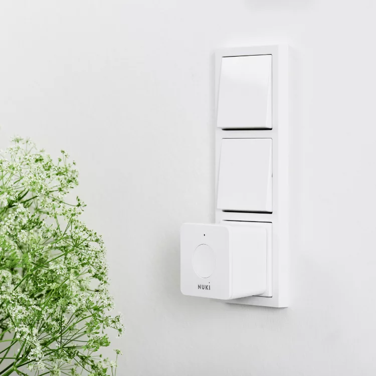 Weiße Wand mit drei Lichtschaltern und einem Nuki Smart Lock. Grüne Pflanze im Vordergrund.