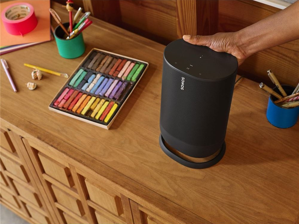 Een zwarte Sonos-luidspreker op een houten kast, met knutselspullen in de buurt. Een hand raakt de luidspreker aan.