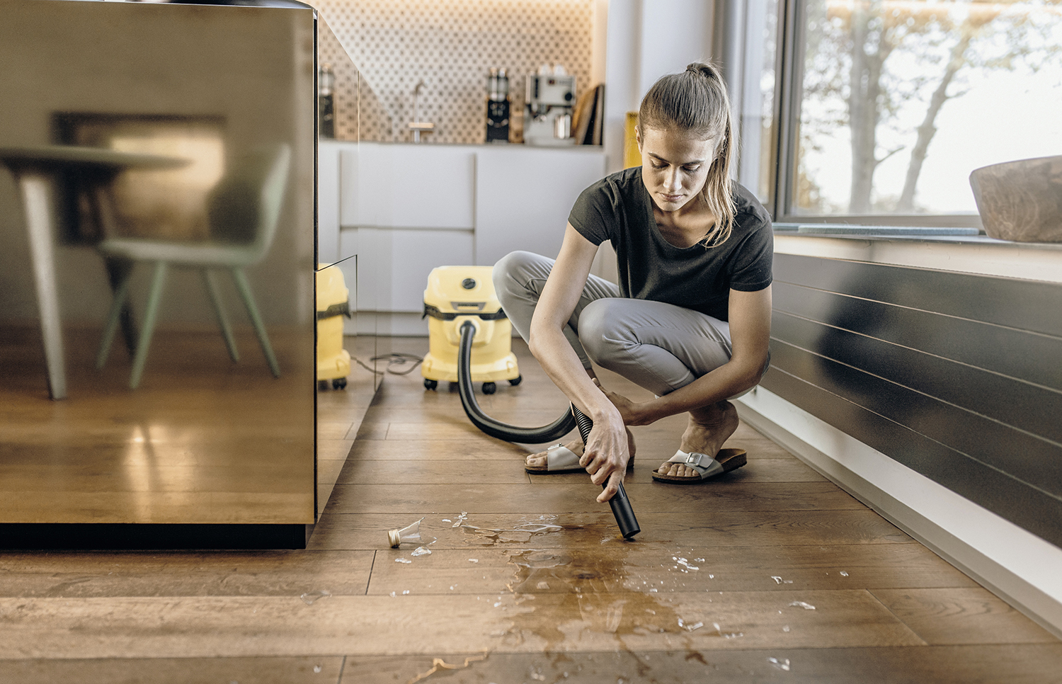 Een vrouw stofzuigt een houten vloer. Water en gebroken glas liggen op de vloer.