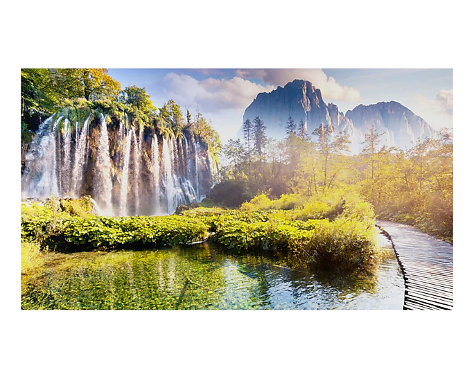 Ein malerischer Wasserfall in einem Wald mit Holzweg. Grünes Laub und ein Berg im Hintergrund.