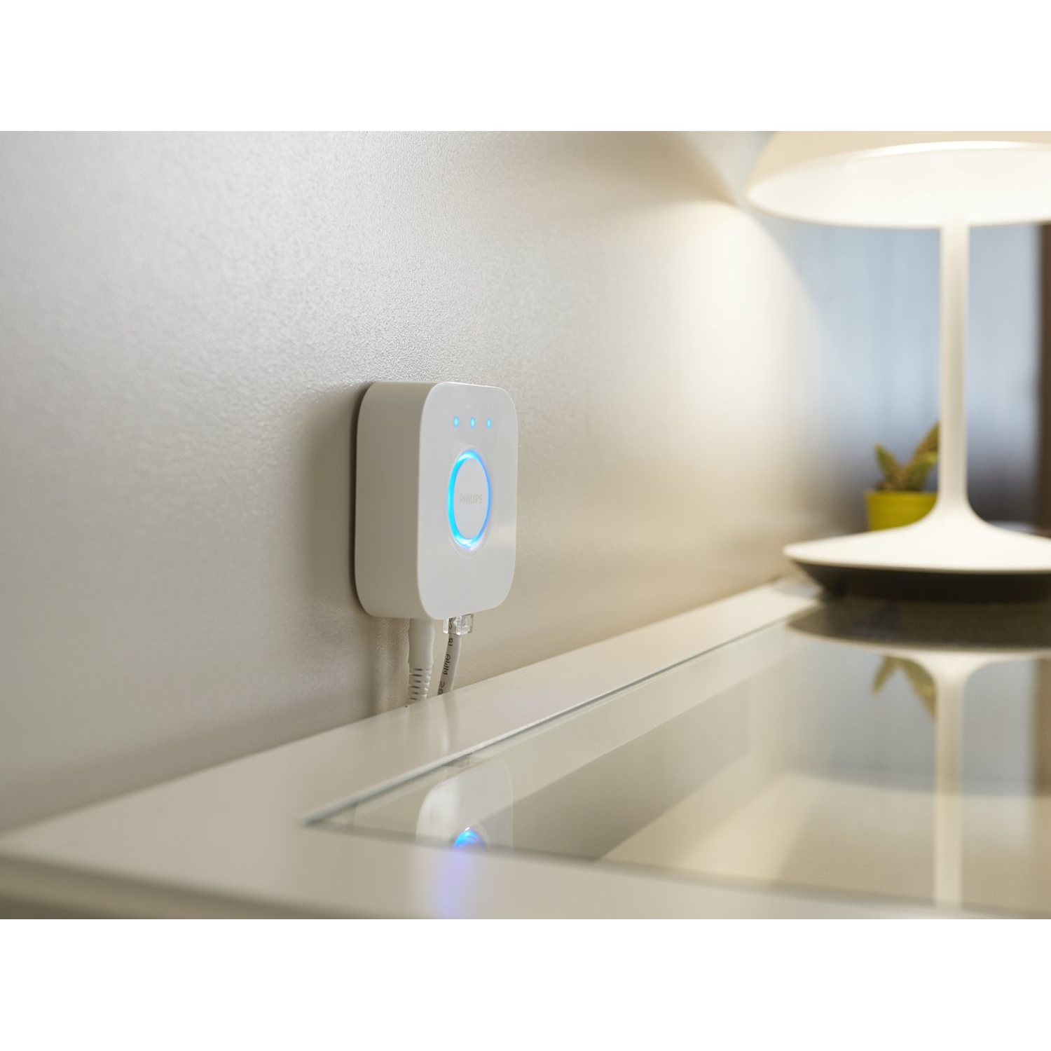 Dispositivo de concentrador de hogar inteligente blanco montado en una pared, con dos cables y una lámpara cerca.