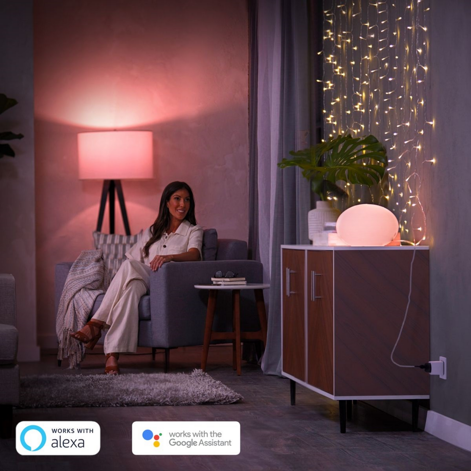 Una mujer sentada en un sillón, iluminada por una lámpara rosa. Un mueble y una guirnalda de luces en el fondo.