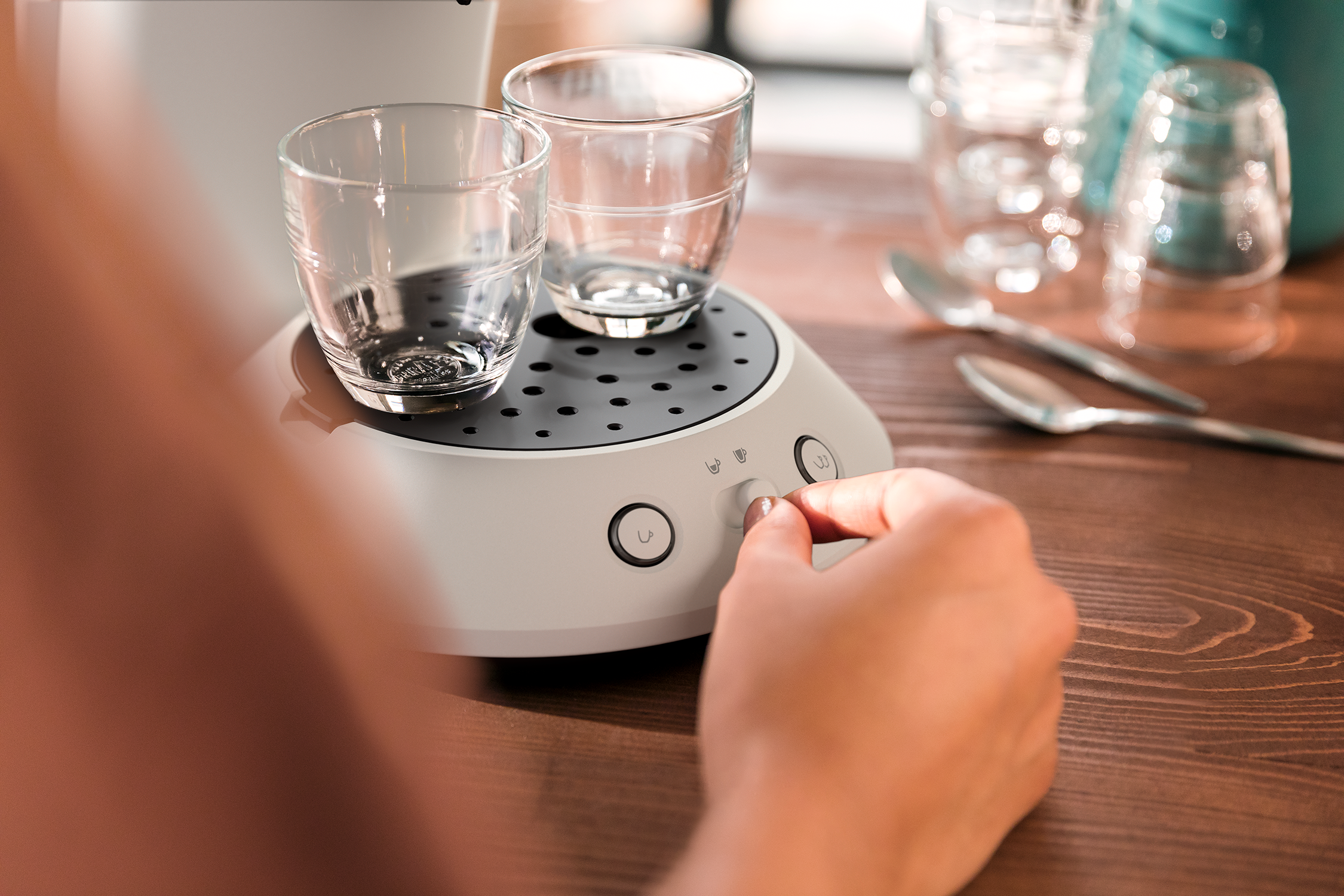 A hand turns a dial on a white device with two glasses. Wooden table in background.