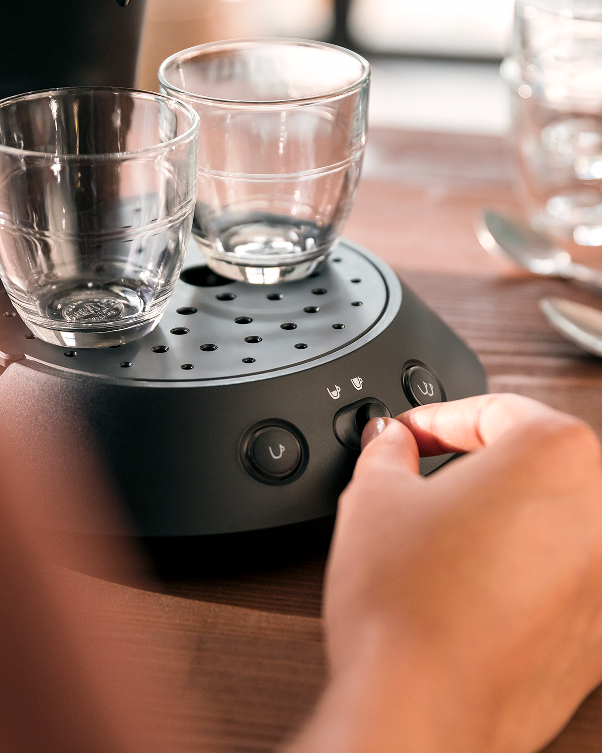 A person's hand adjusting the setting on a coffee maker. Two glasses are on the machine.