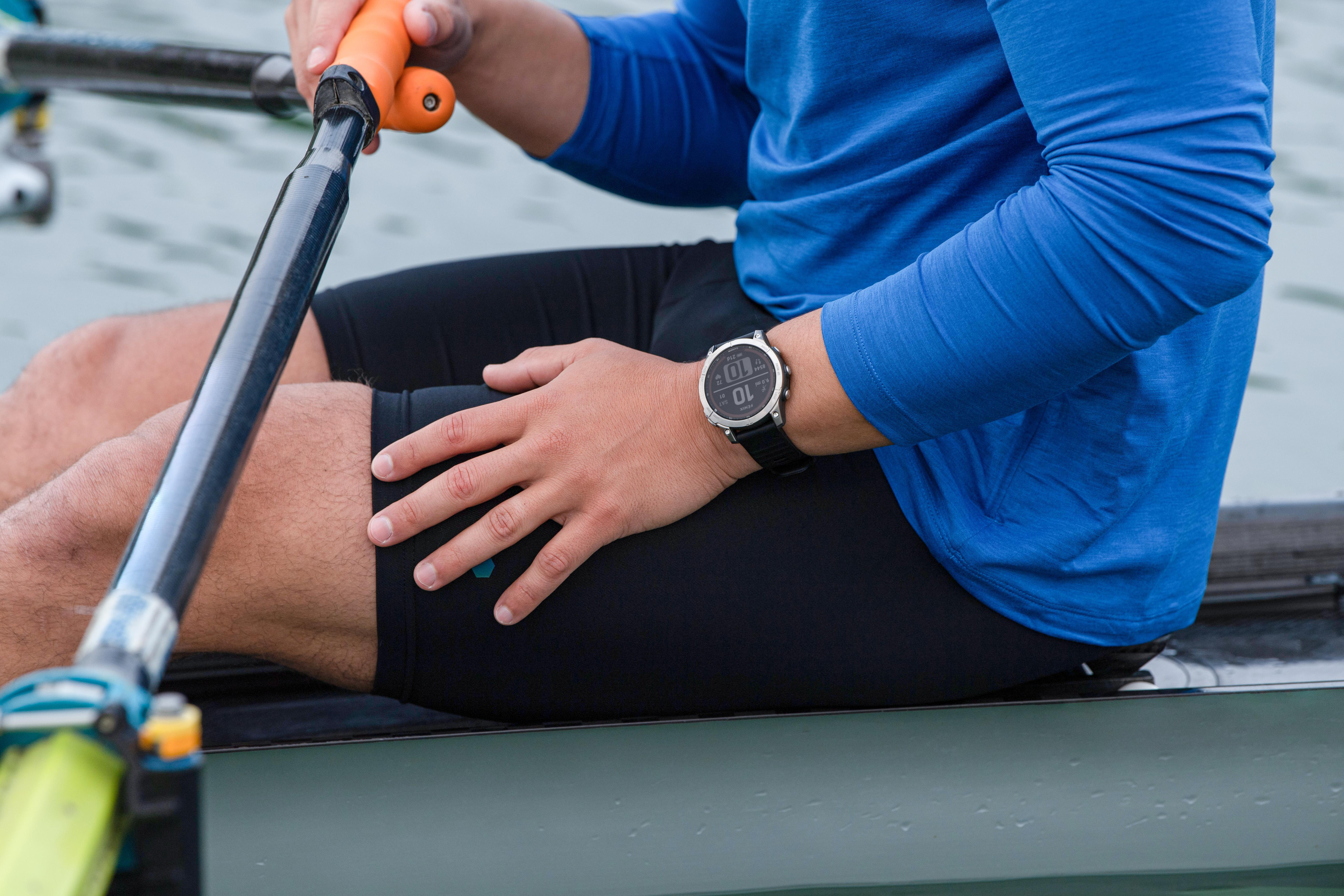 Person mit Smartwatch beim Rudern, blaues Hemd und schwarze Shorts. Wasser im Hintergrund.