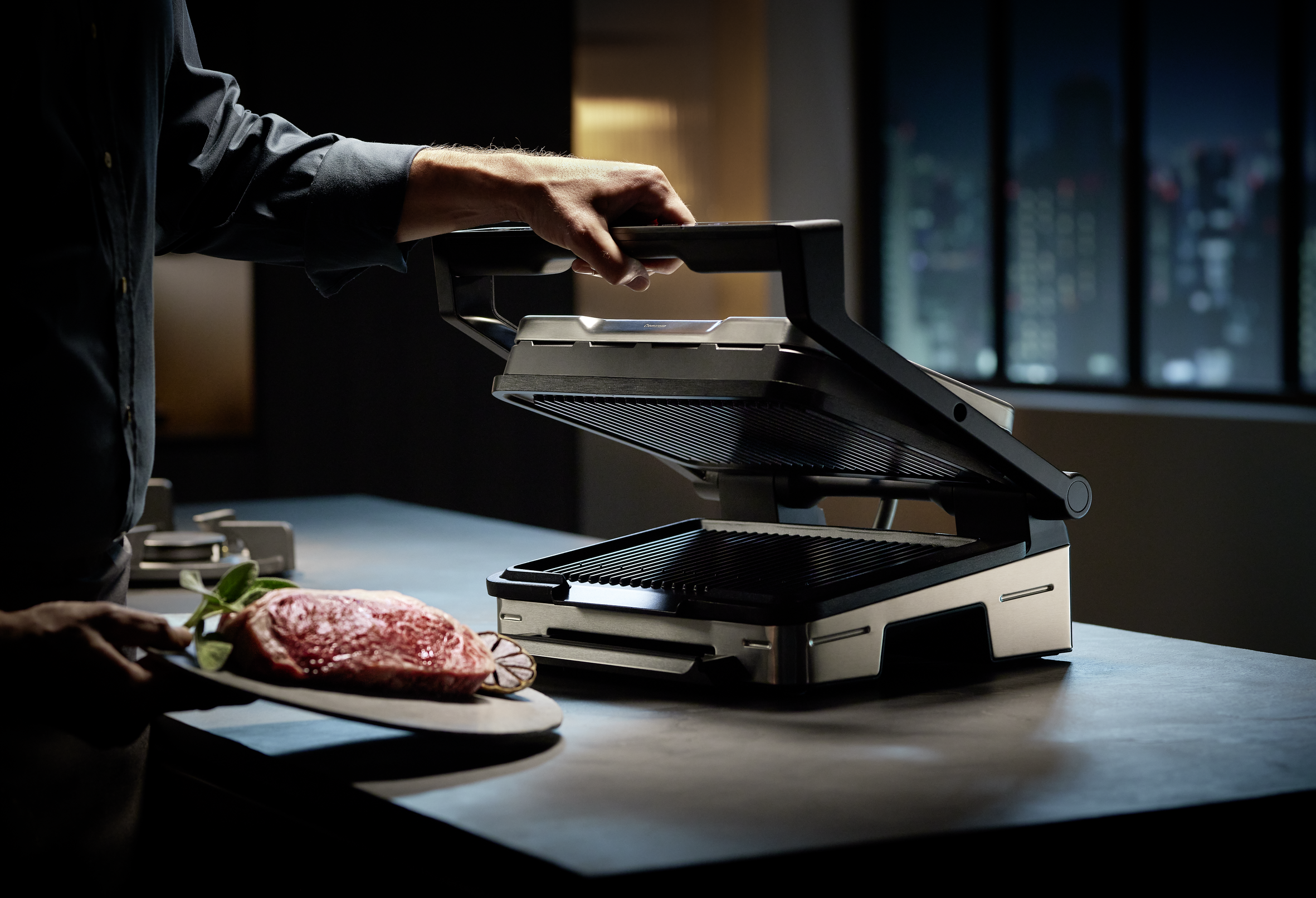 Eine Person grillt ein Steak mit einem geschlossenen Grill. Ein Teller mit Steak ist im Vordergrund.
