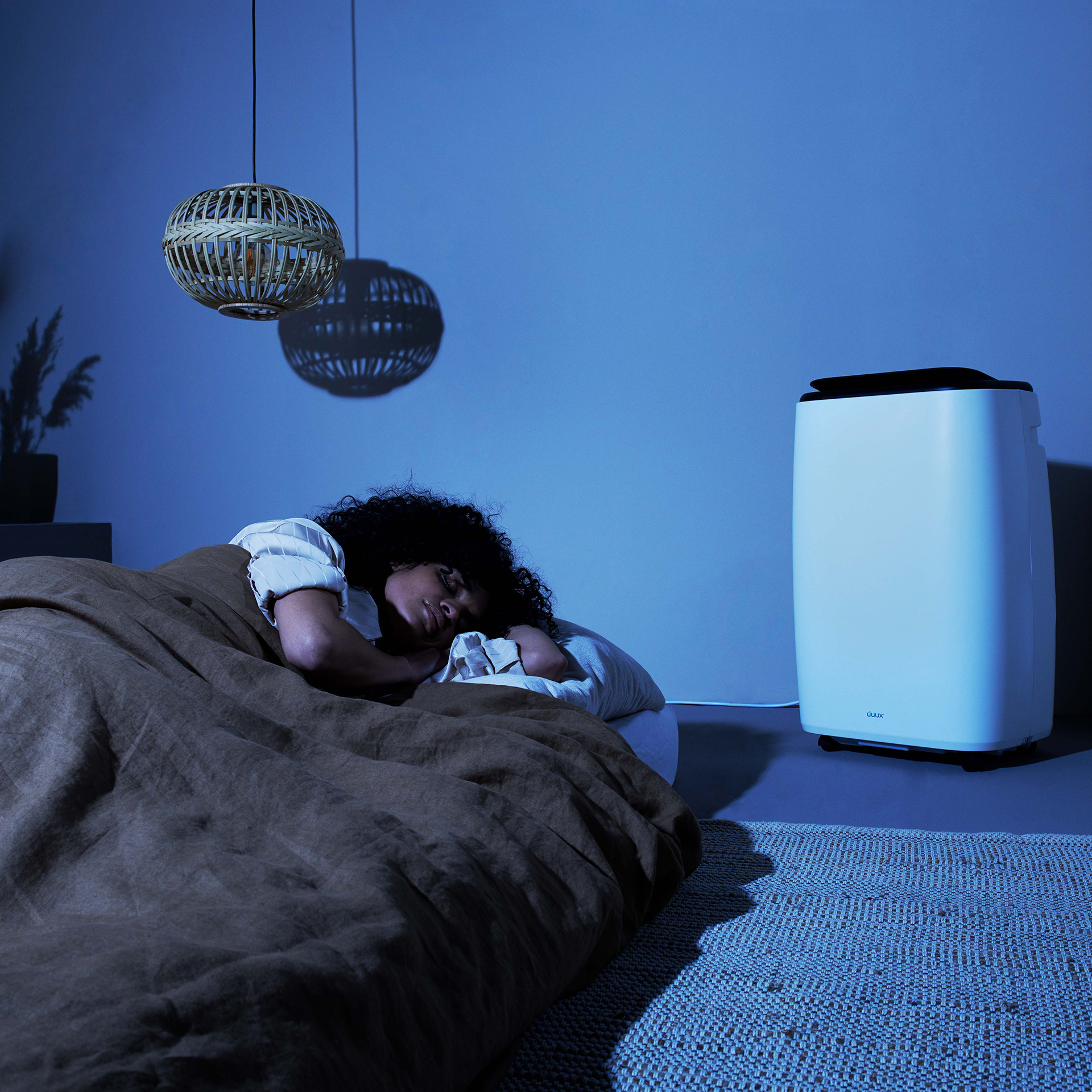 Une personne dort dans un lit. Un purificateur d'air est à proximité. Deux lampes pendent du plafond. Un tapis est au sol.