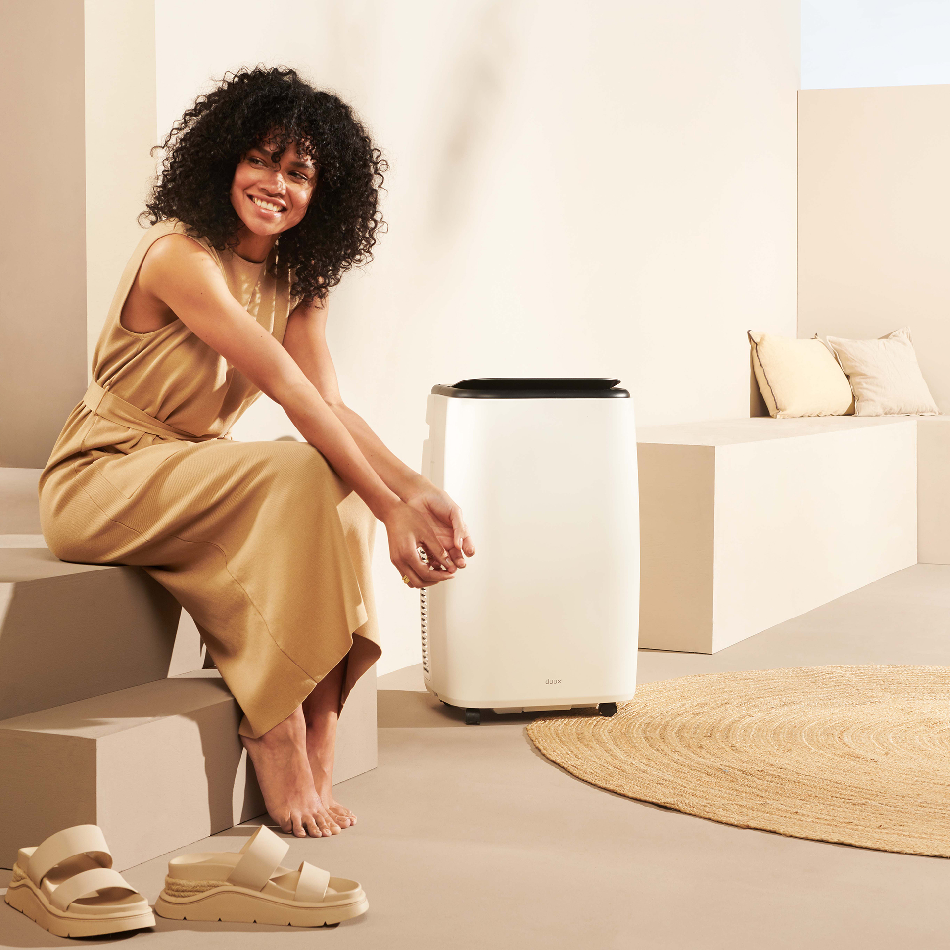 Une femme sourit près d'un purificateur d'air blanc. Elle est assise sur des marches. Des sandales sont au sol. Un tapis et des oreillers sont visibles.