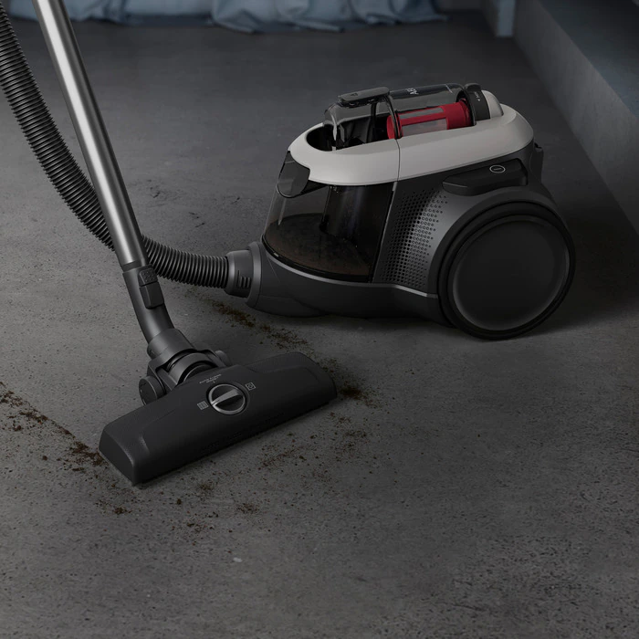 Black and white vacuum cleaner on gray floor. Hose and brush visible. Dust and debris are present.