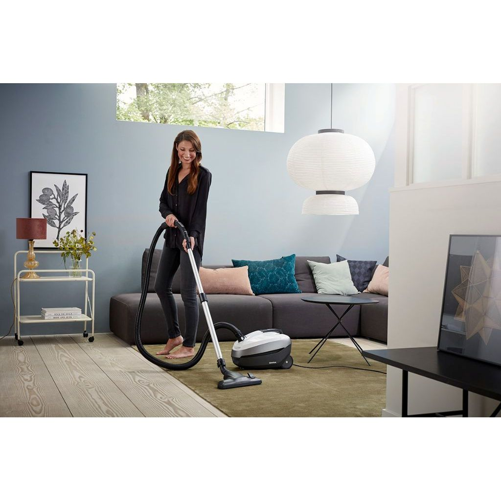 Woman vacuuming a room with a gray sofa and a white pendant lamp. Wooden floor.