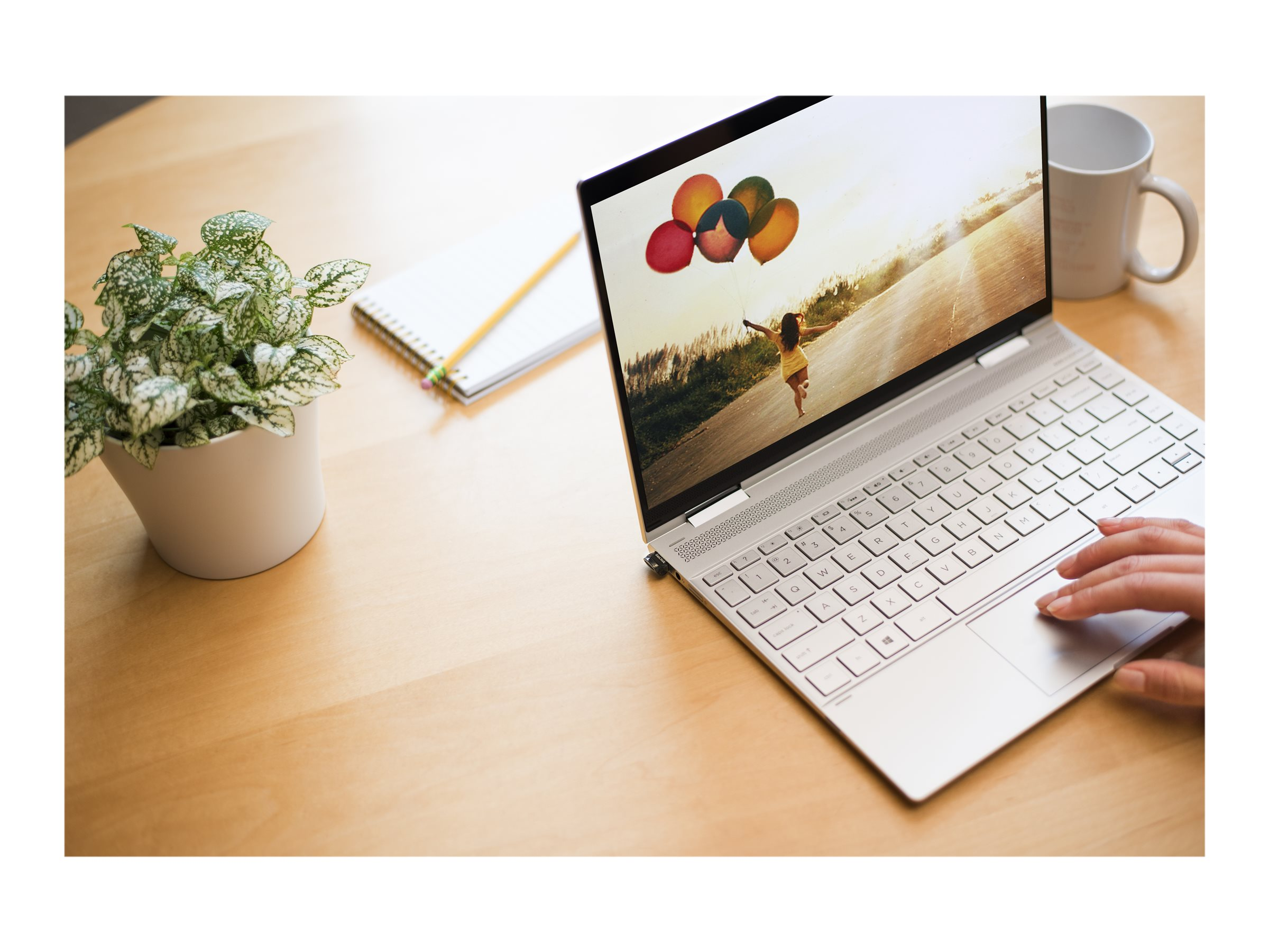 Laptop auf Holztisch. Bildschirm zeigt Kind mit Ballons. Pflanze, Notizblock und Tasse sind ebenfalls vorhanden.
