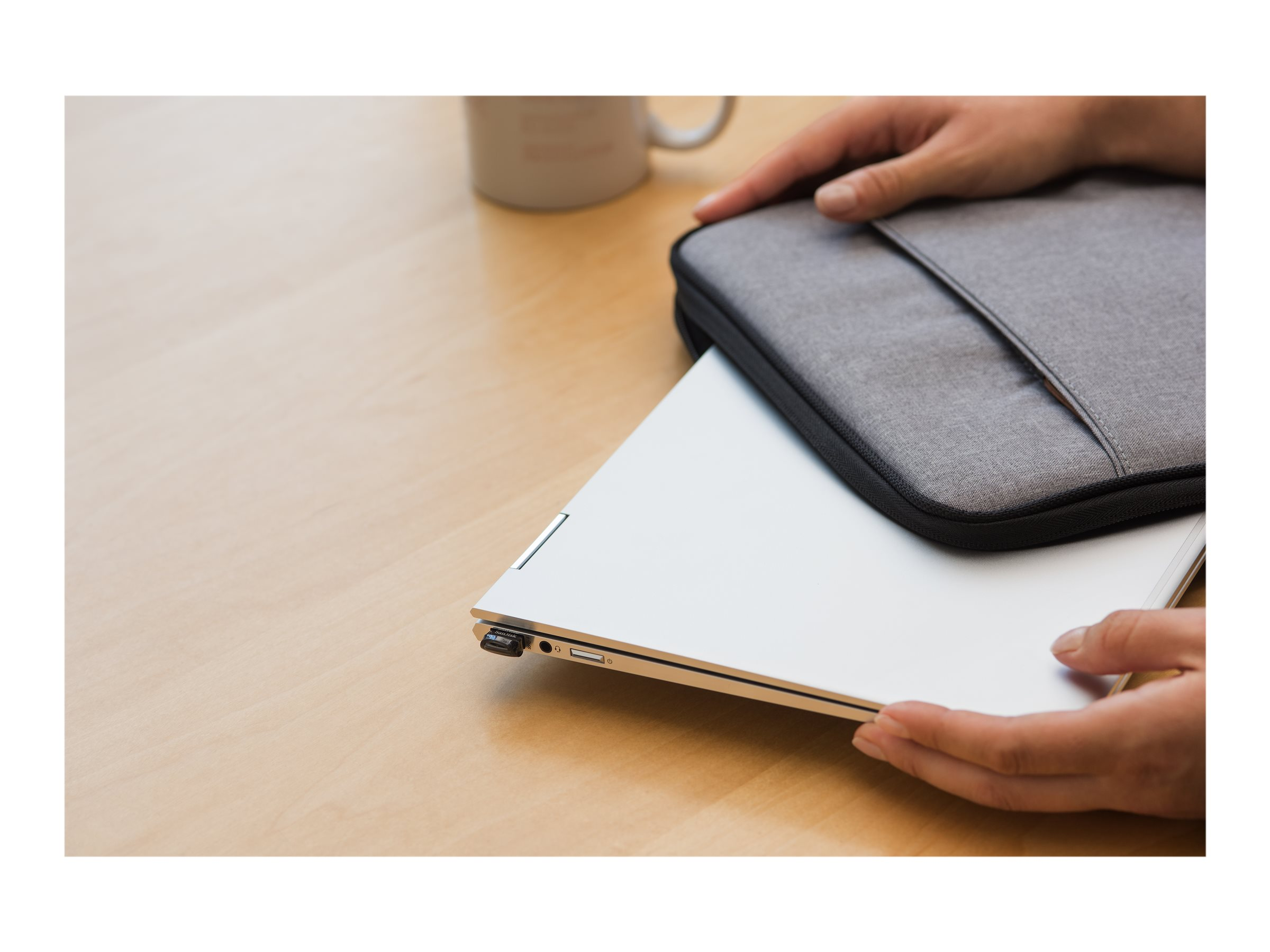 Hände halten einen silbernen Laptop neben einer grauen Laptoptasche auf einem Holztisch.
