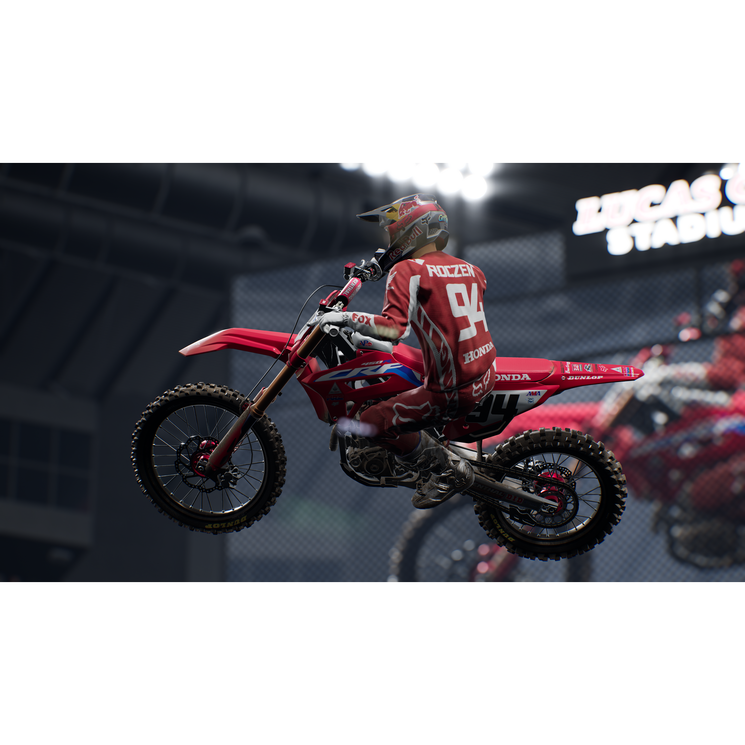 Un piloto de motocross rojo y blanco en el aire. El estadio está al fondo.