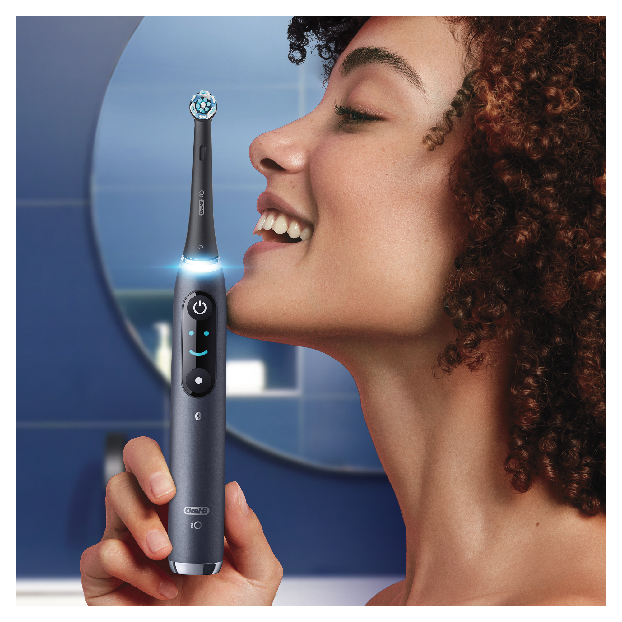 Mosolygó nő, szürke elektromos fogkefét használ. Egy tükör a háttérben.