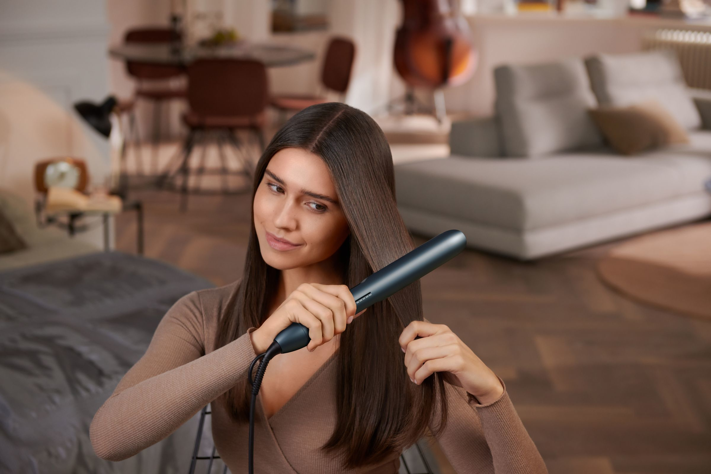 Femme se lissant les cheveux avec un fer plat, près du visage, pull beige, fond flou.