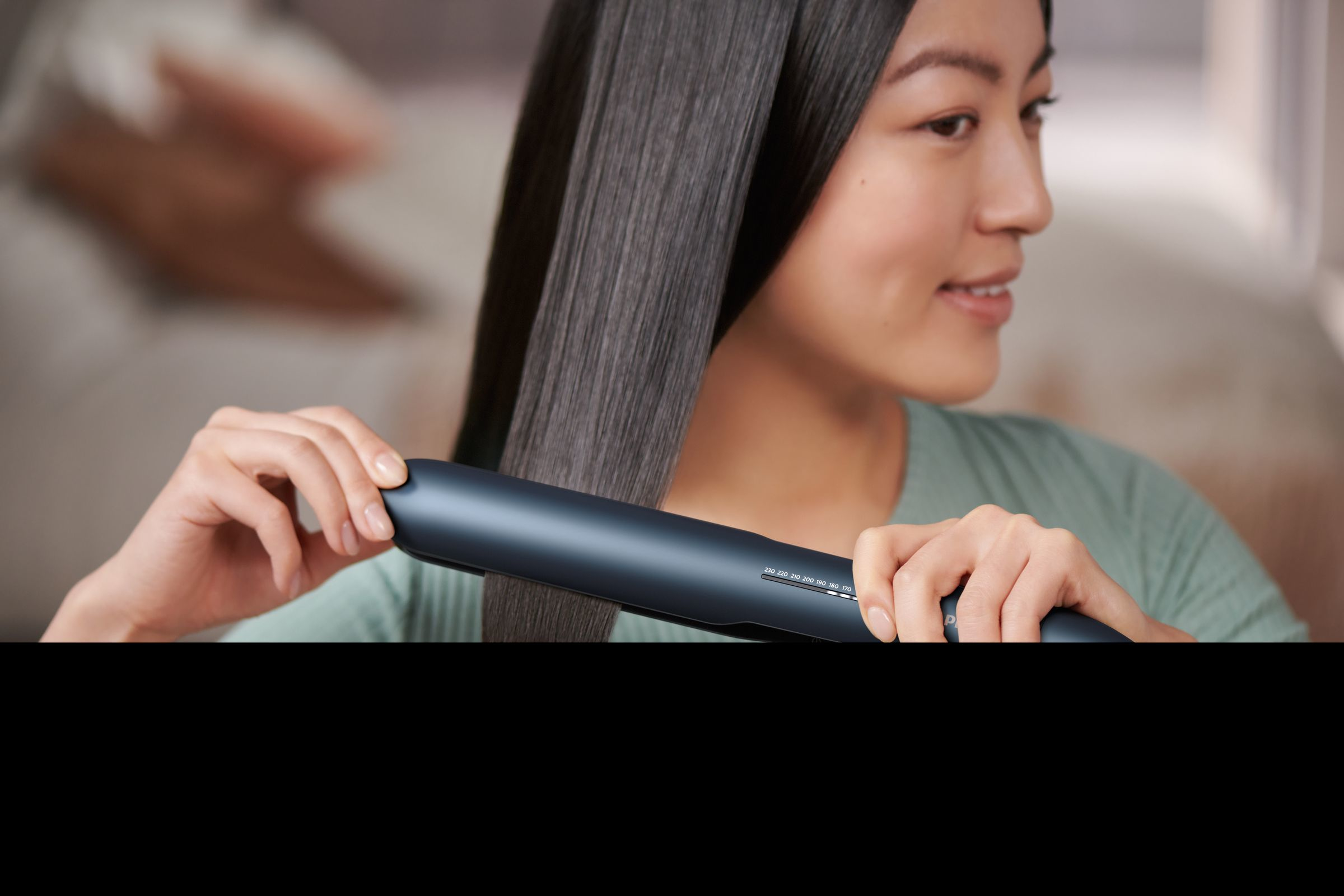 Une femme utilise un lisseur de cheveux de couleur foncée pour lisser ses cheveux, avec un arrière-plan flou.