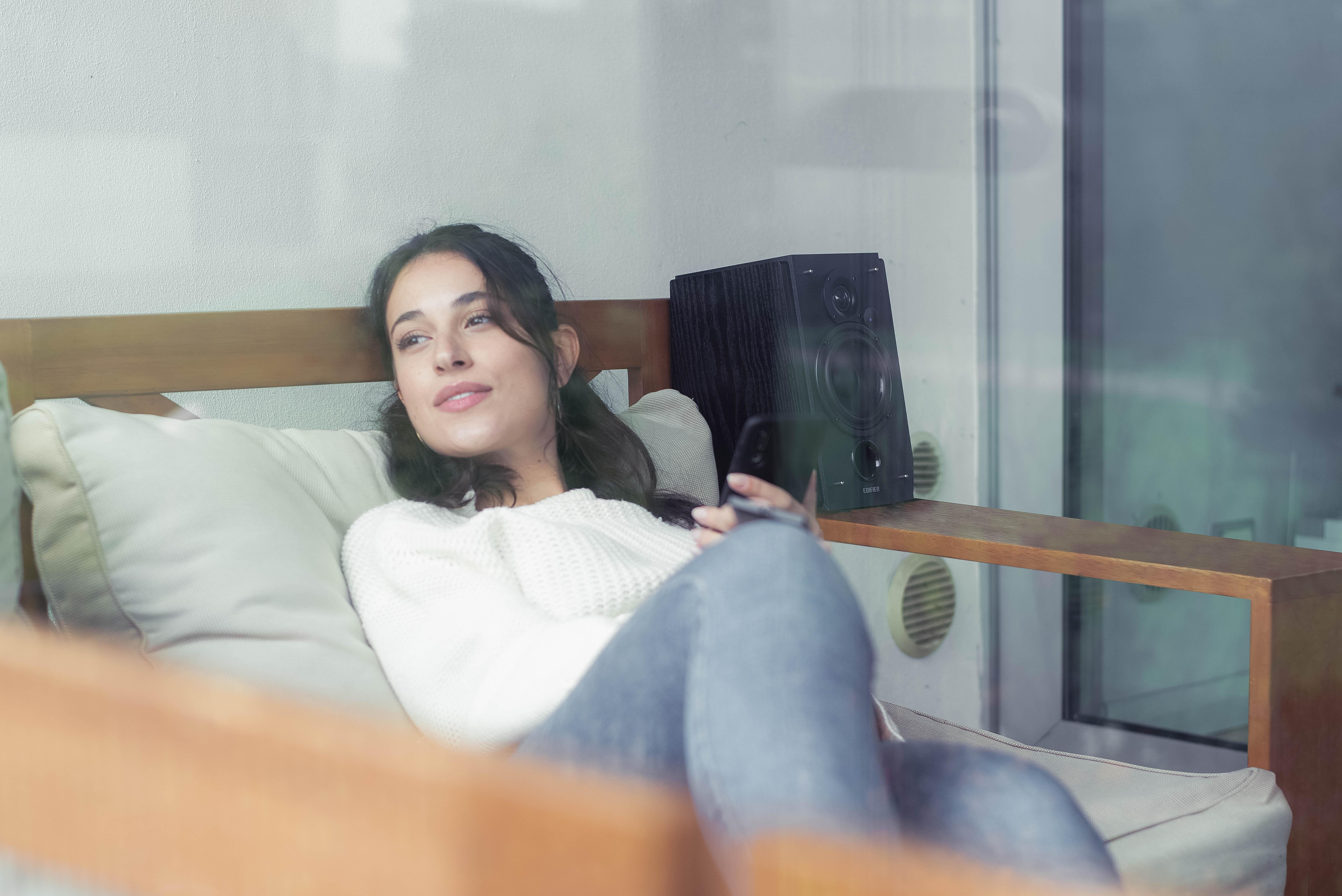 Frau auf Sofa mit Lautsprecher. Hält Fernbedienung. Fenster im Hintergrund.