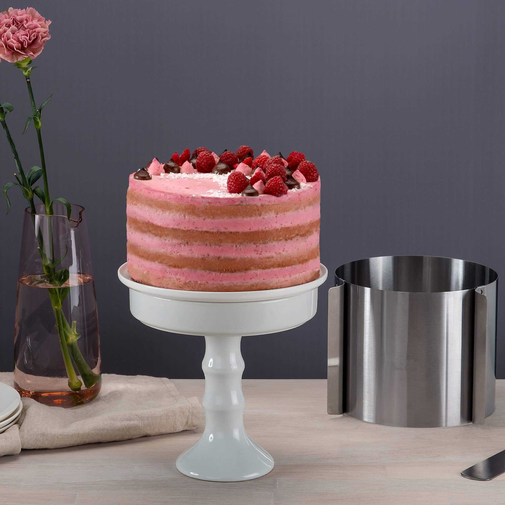 Rosa geschichteter Kuchen auf einem weißen Ständer. Rosa Blume in einer Vase links. Ein Metallring rechts.