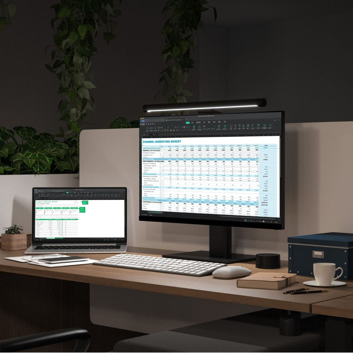 Irodai beállítás laptop-pal és monitorral. A monitor egy táblázatot mutat. Asztalon lámpa és kék doboz.