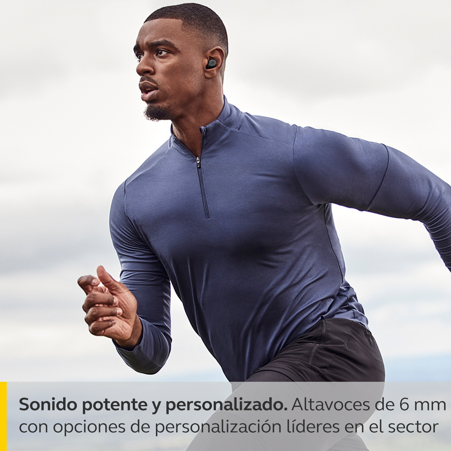 Hombre corriendo con auriculares negros. Camisa azul, shorts negros, fondo nublado.