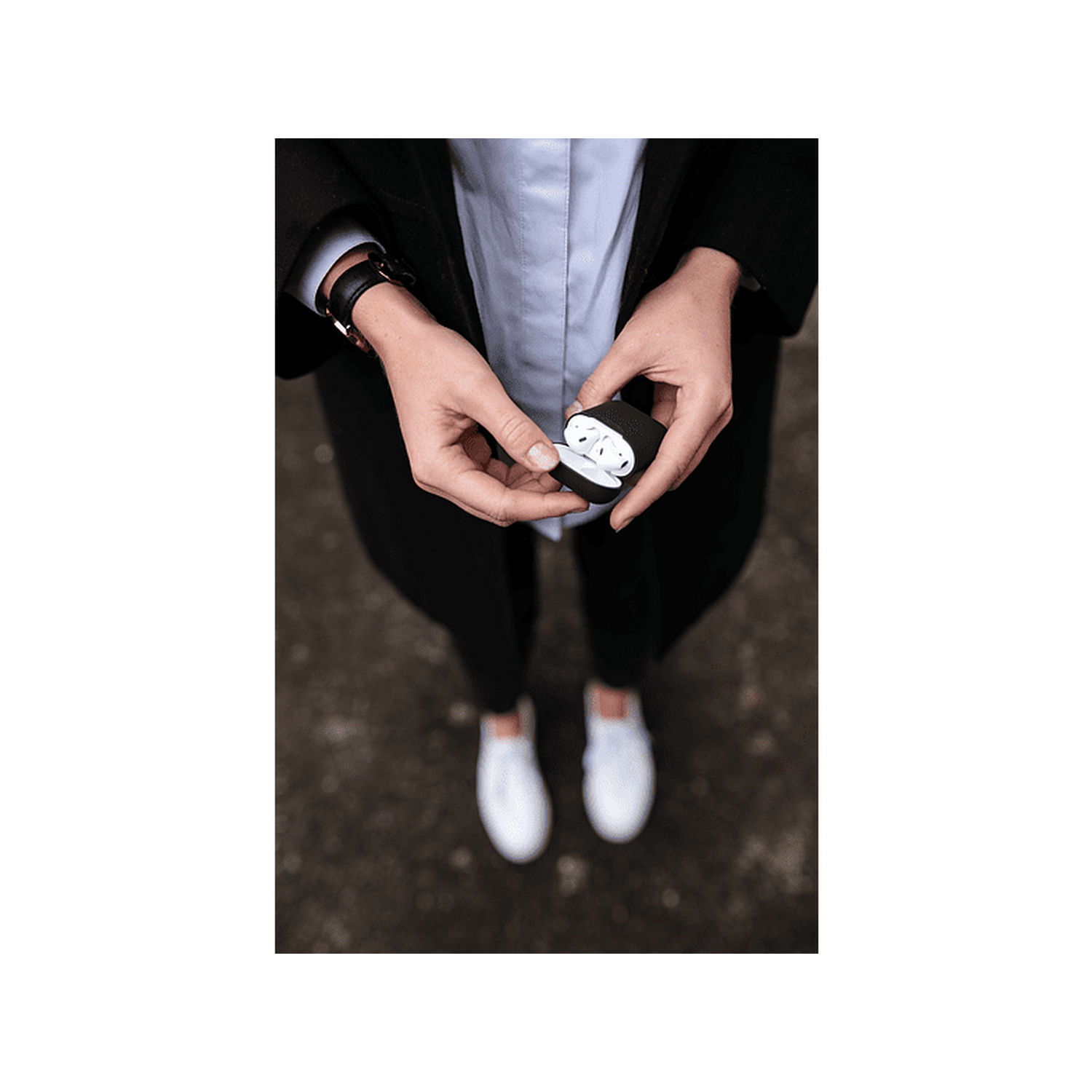 Una persona sostiene un estuche abierto con auriculares blancos. Lleva un abrigo negro y pantalones negros.