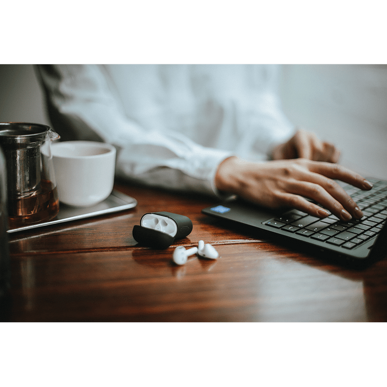Una persona trabajando en una laptop con una taza y unos auriculares blancos en la mesa.