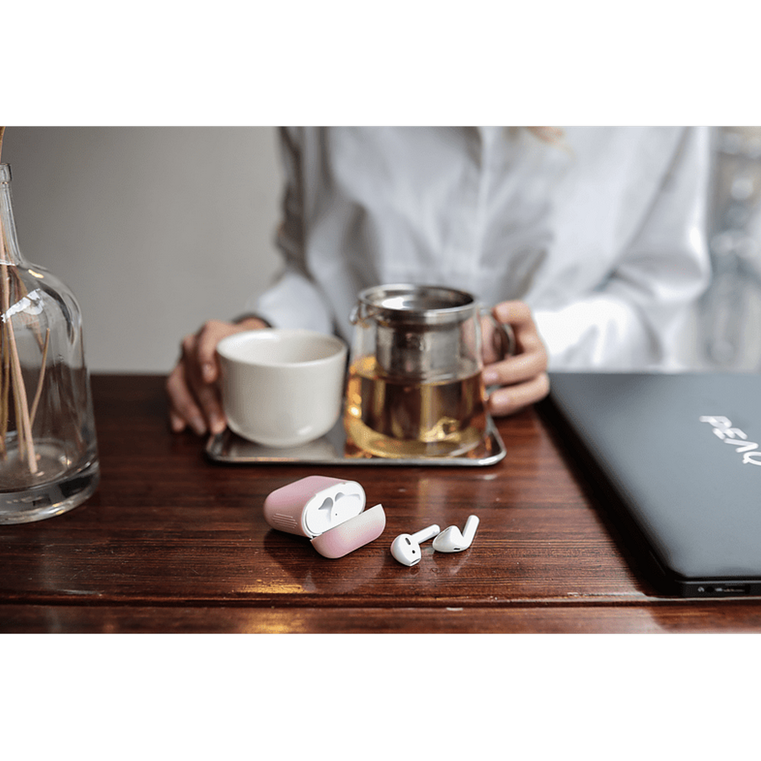 Auriculares en un estuche rosa sobre una mesa, junto a un juego de té y un portátil.