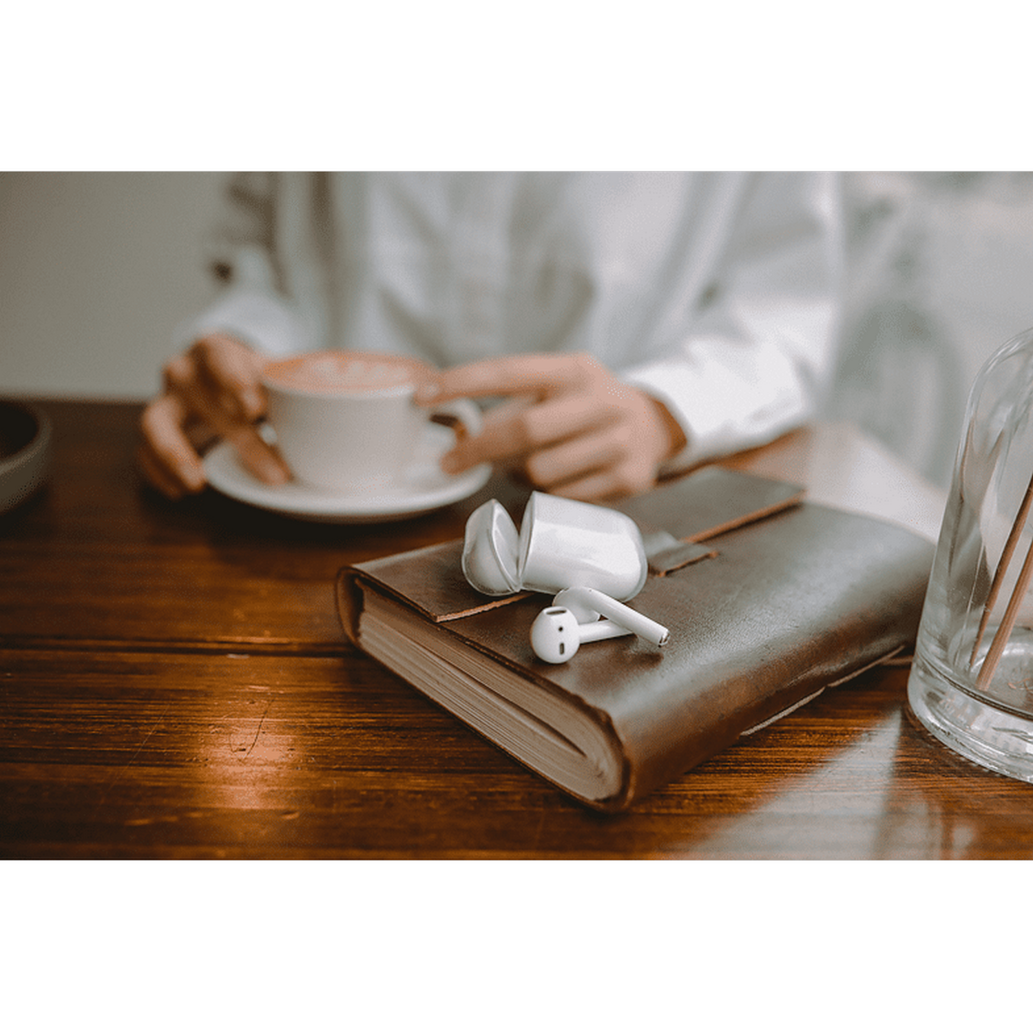 Una persona sosteniendo una taza cerca de un cuaderno de cuero marrón con auriculares y un tarro de cristal.