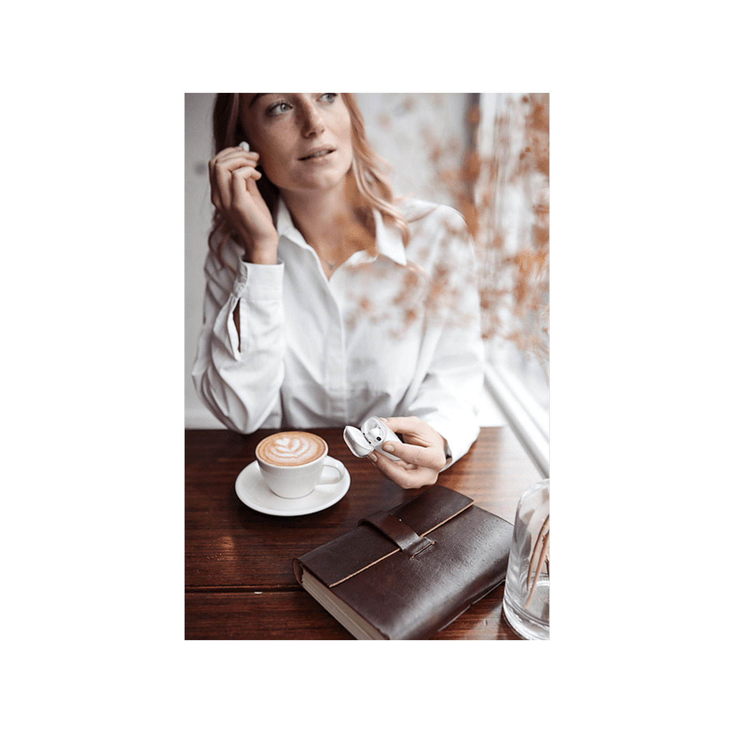 Mujer en mesa con café, diario y auriculares. Sostiene un auricular, mirando hacia arriba y sonriendo.