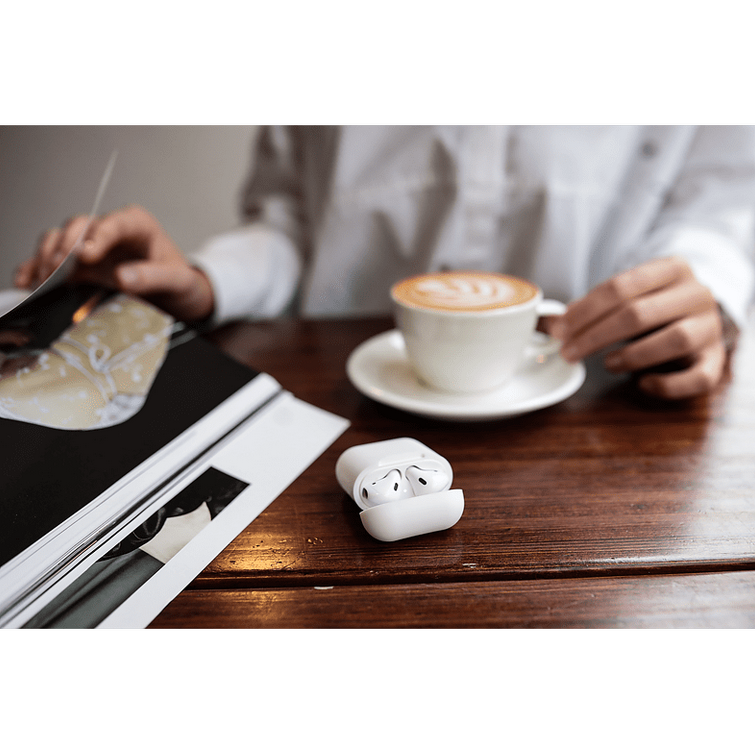 Estuche blanco de AirPods con AirPods junto a una taza de café y una revista en una mesa de madera.