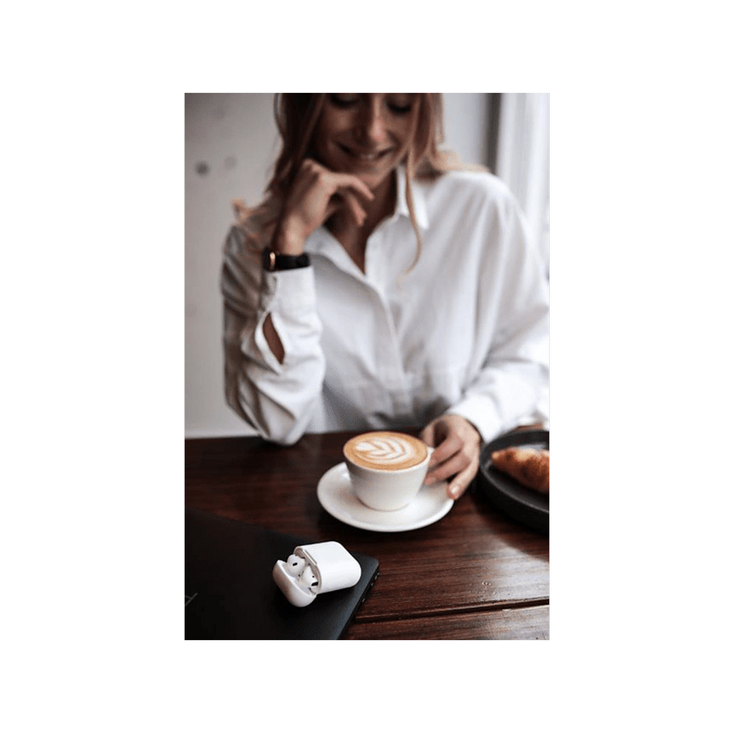 Una mujer con una camisa blanca sostiene una taza de café con arte latte. Auriculares blancos al lado.