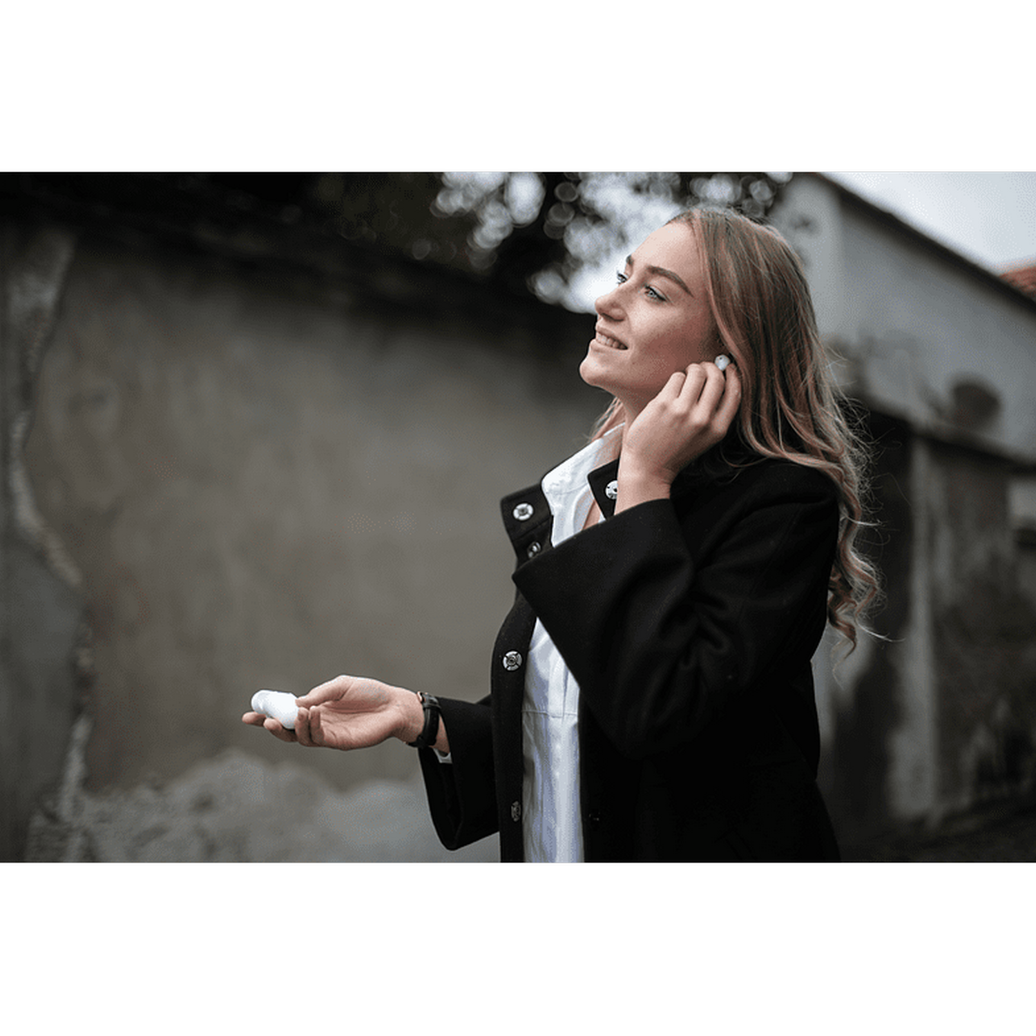 Una mujer sosteniendo un auricular blanco. Lleva un abrigo negro y sonríe. El fondo está borroso.