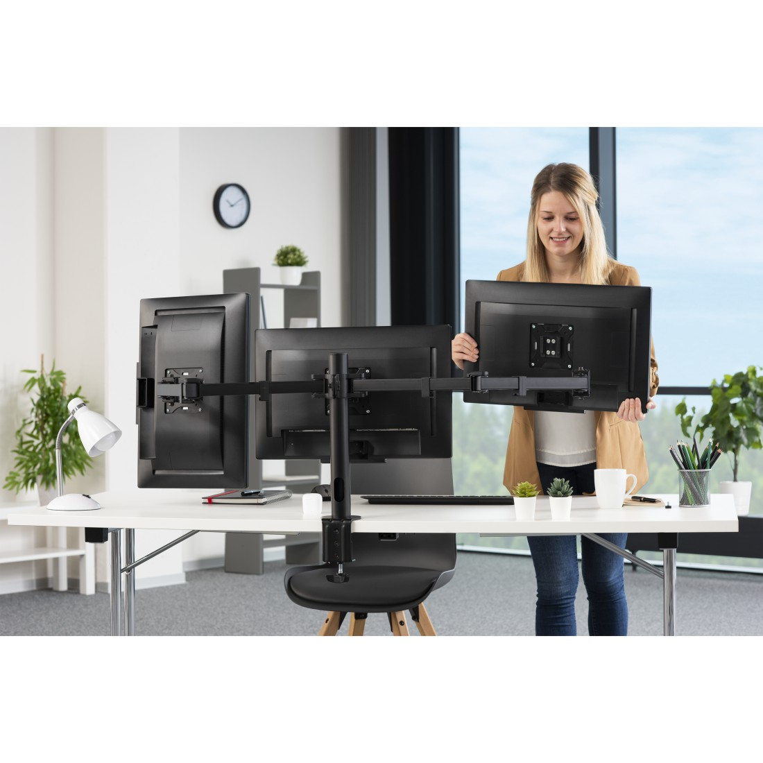 Frau hält einen Monitor in einem Büroumfeld mit zwei weiteren Monitoren auf einem Schreibtisch.